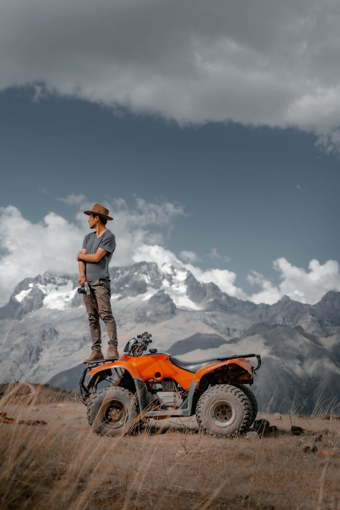 A man in a hat stands on a quad, set against Peru's dramatic Cusco mountains. Adventure and exploration.